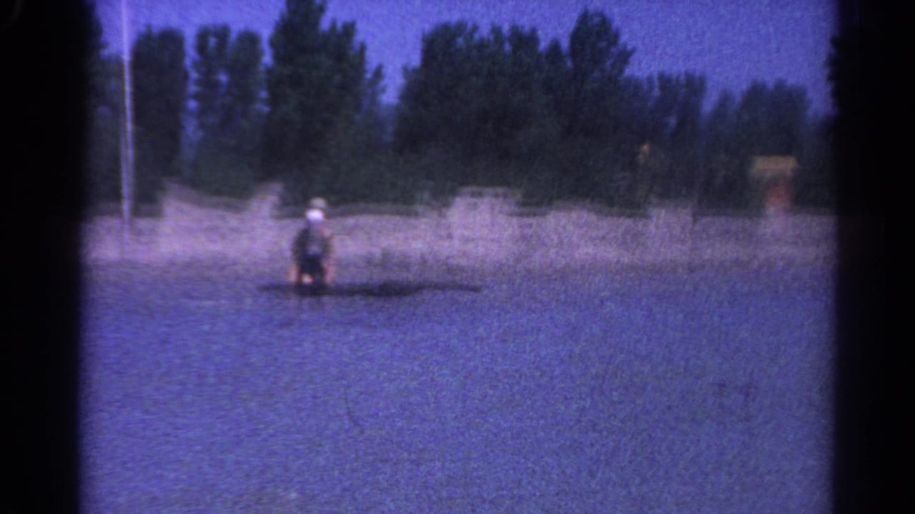 Woman stands or kneels on a small board in the lake with rippled water and trees behind her.
