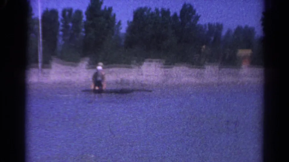 The woman remains on the small board in shallow Catskill lake water, framed against a sandy beach and dense trees.