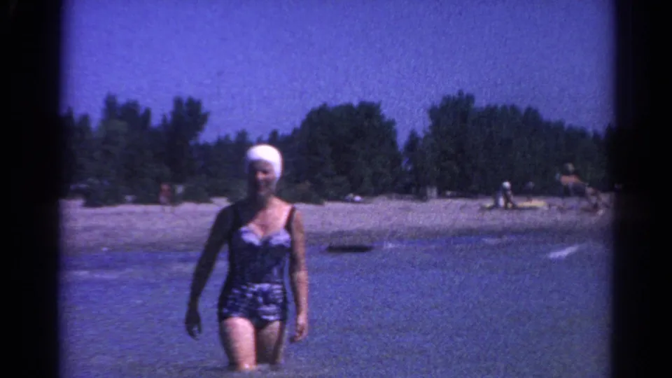 A woman in a blue one-piece swimsuit and white swim cap walks through shallow lake water at Catskill after the surfing attempt.