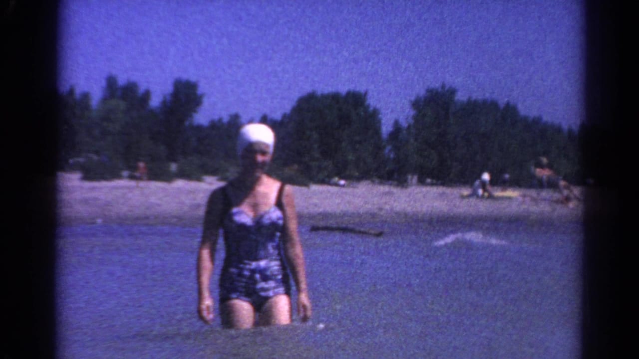 In 1962 Catskill, New York footage, a woman in a patterned swimsuit and white swim cap walks through shallow lake water near a sandy, tree-lined shore.