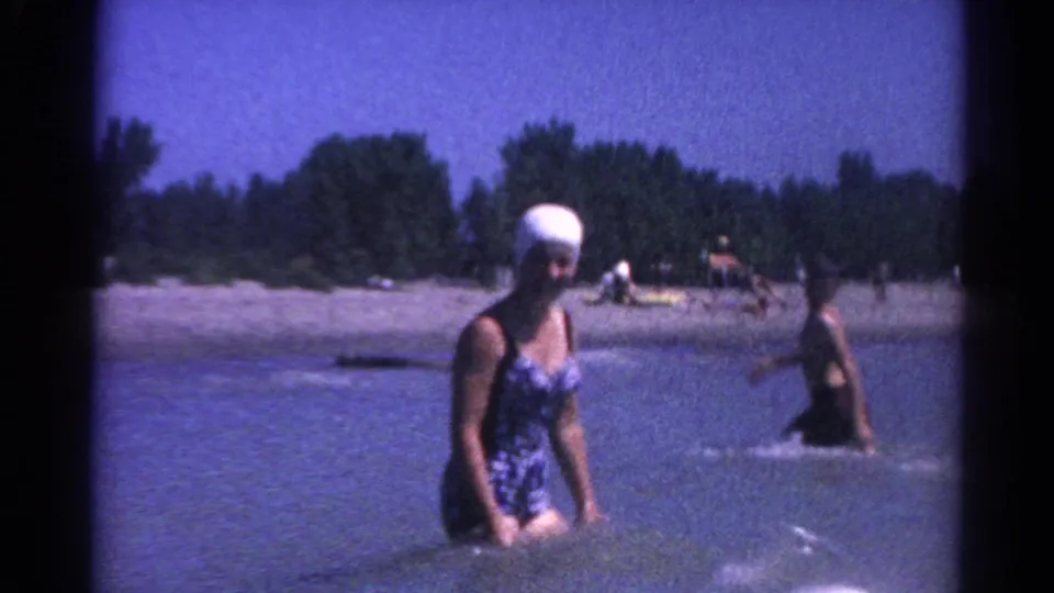 A woman in a white swim cap and one-piece swimsuit wades in the lake as another swimmer splashes in the background near the beach.
