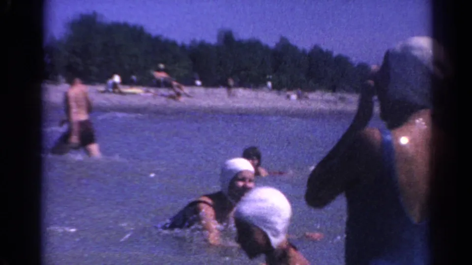 Multiple swimmers, including figures in white swim caps, play and splash in shallow lake water with beachgoers and trees behind them.