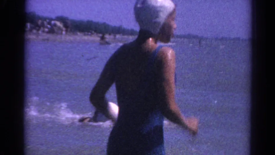 A woman in a white swim cap and dark swimsuit turns in shallow water while other swimmers splash farther out in the lake.