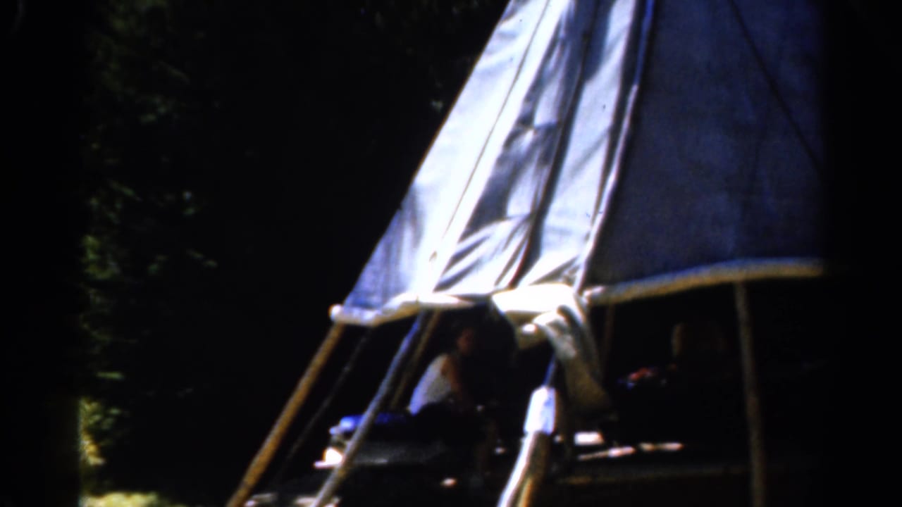 Canvas tent in a shaded wooded campsite with seated figures visible inside.