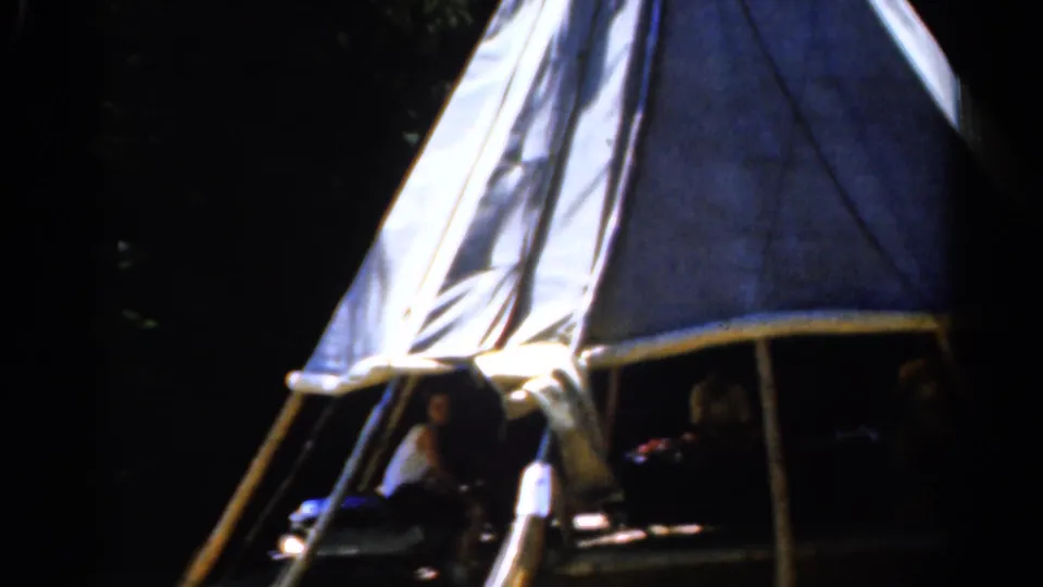 1962 Catskill, New York frame showing the tent entrance and shadowed campers, with bright canvas and wooded background.