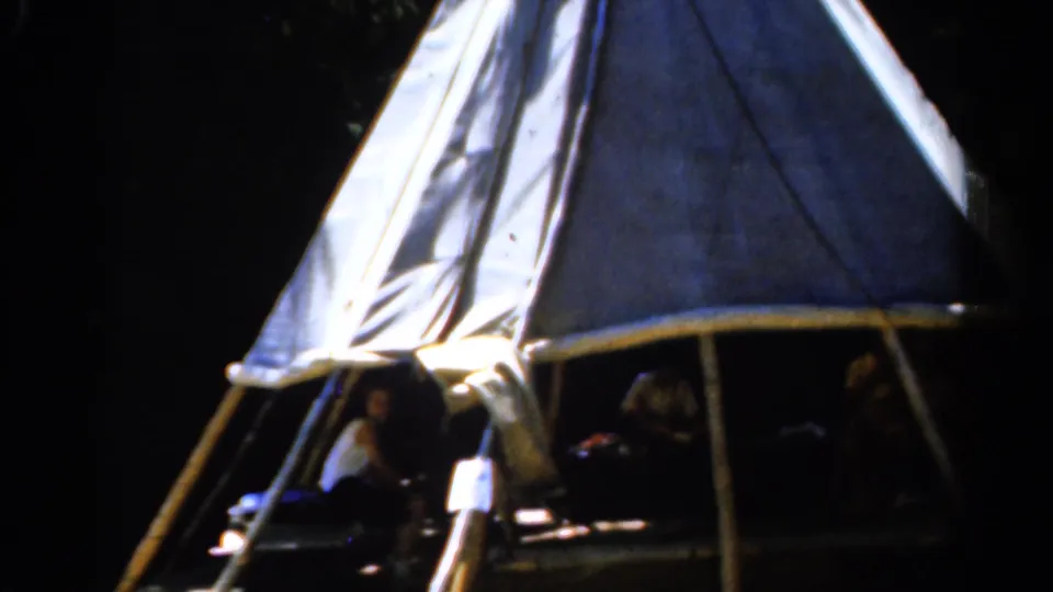 1962 Catskill, New York home-movie frame of a wooded campsite, canvas tent, and family members seated beneath the awning.