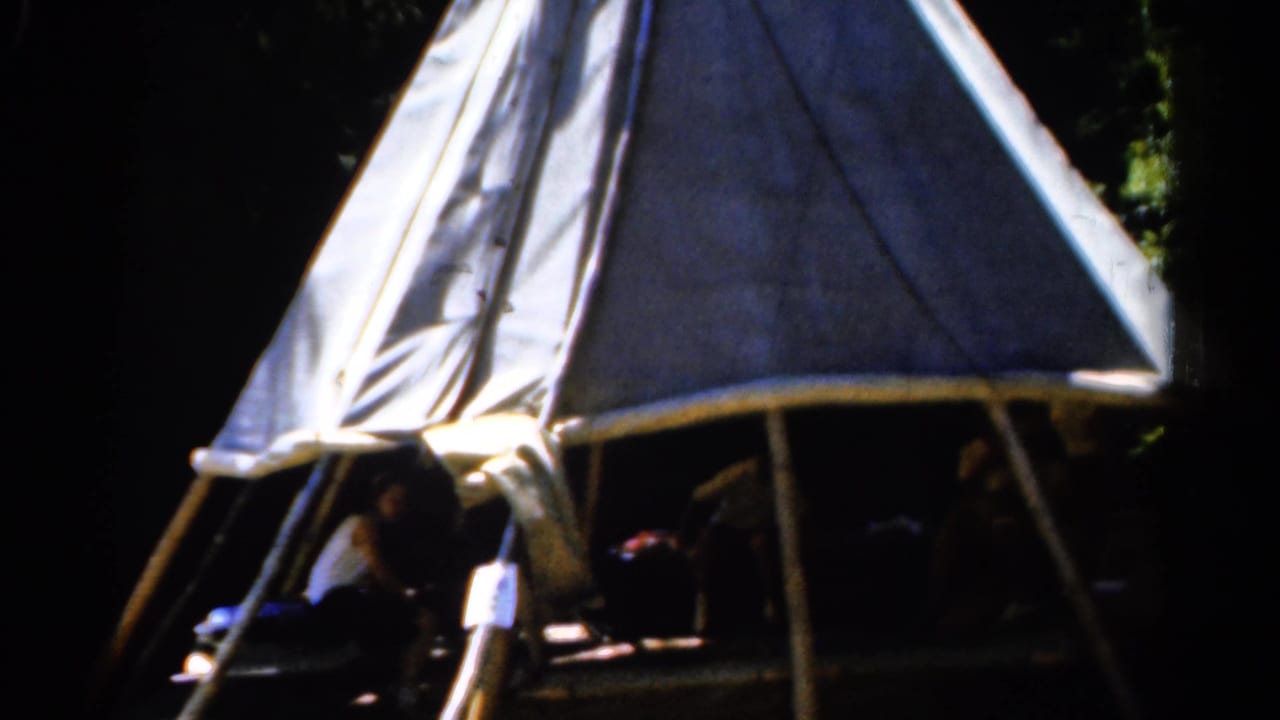 1962 Catskill frame of a bright canvas teepee-like structure with wooden poles and several people obscured by the shaded interior.