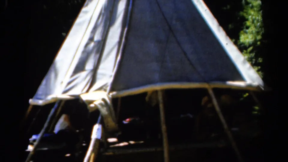 1962 Catskill home-movie frame showing the teepee-like shelter closer in view, with one seated person near the entrance and others in deep shade.