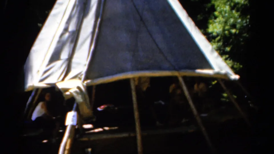 1962 Catskill frame of a sunlit canvas shelter set against trees, with several people faintly visible under the roofline.