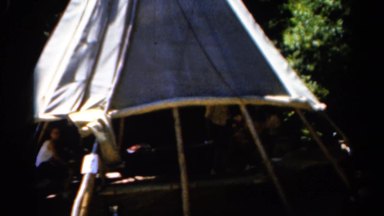 Canvas tent with seated people in shadow and green trees behind it.
