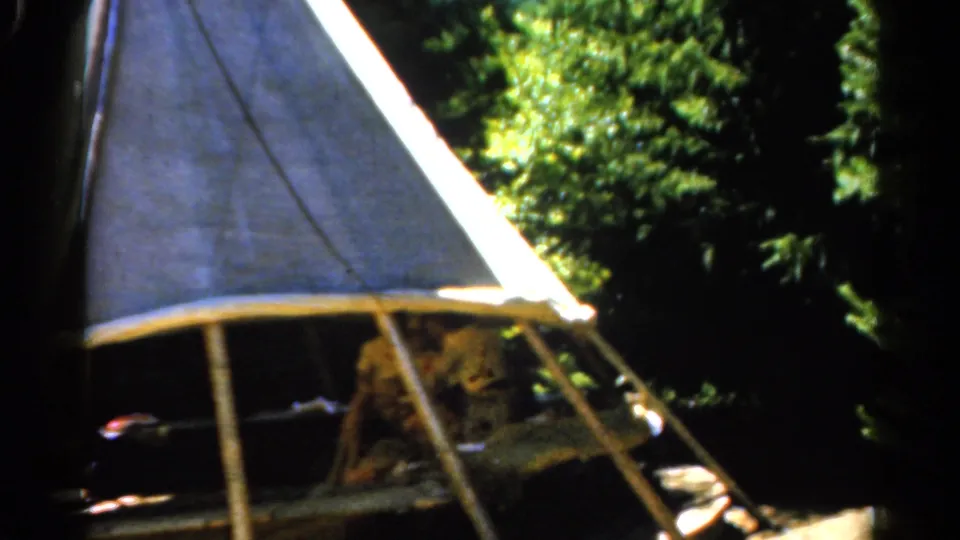 1962 Catskill home-movie frame showing a person shifting inside a shaded canvas tent beside bright green trees.
