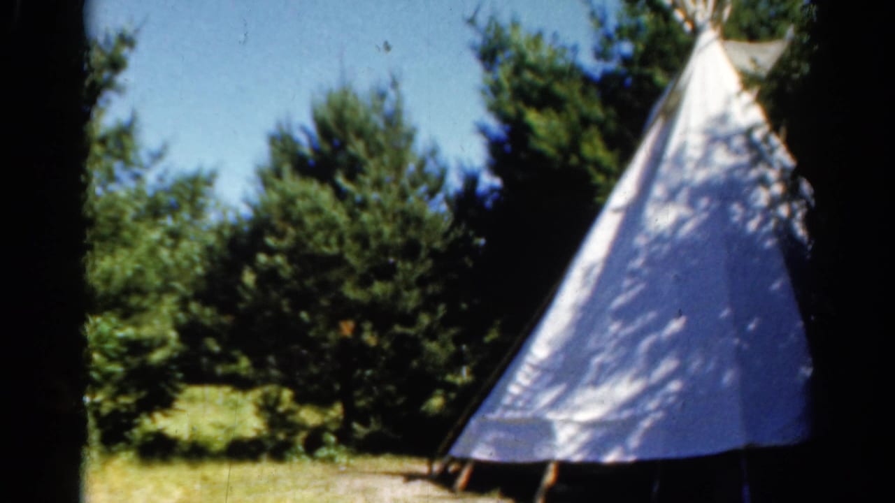1962 Catskill home-movie frame of a white teepee-style camping tent set against pine trees and blue sky.