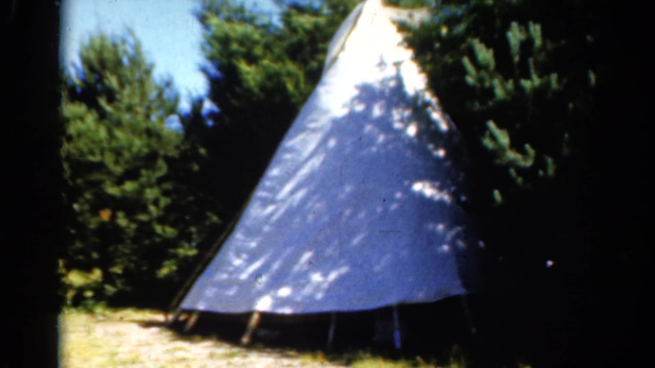 White teepee-style canvas shelter among trees at a wooded campsite.