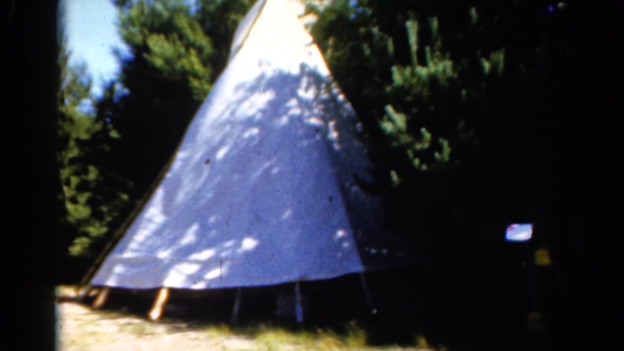 1962 Catskill 8mm frame of a teepee-style canvas shelter beside evergreens, with a small sign or marker visible at right.