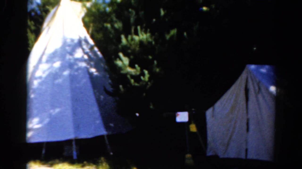 1962 Catskill home-movie frame showing two pale tents beneath dense trees with dappled sunlight and dark shadow.