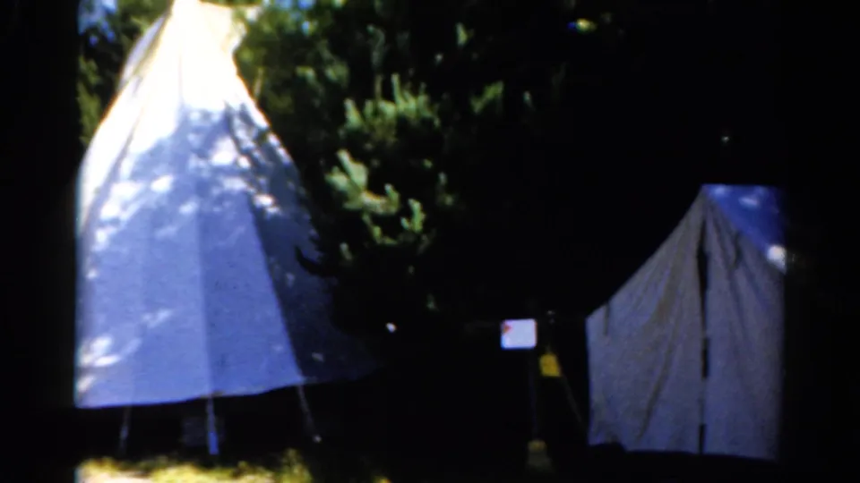 1962 Catskill home-movie frame showing two pale tents beneath dense trees with dappled sunlight and dark shadow.