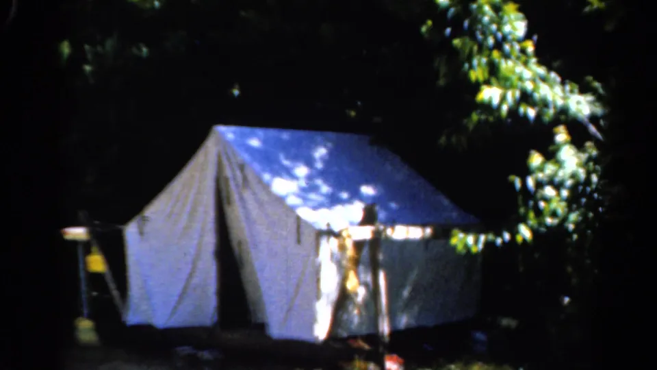 1962 Catskill camping footage showing a white canvas tent under dense trees with sunlight across the roof and front wall.