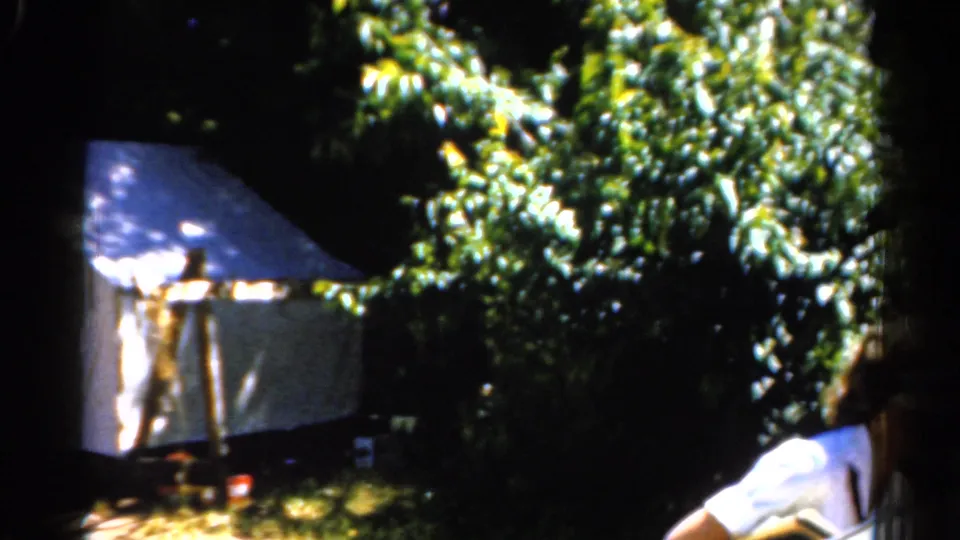 1962 Catskill home-movie frame with a woman in a white shirt holding a stringed instrument near a tent hidden by foliage.