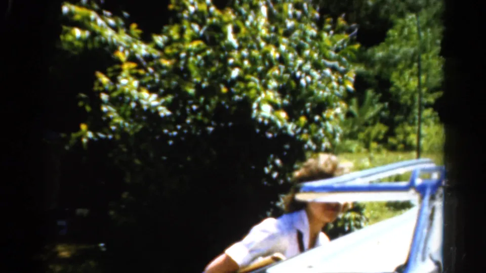 1962 Catskills home-movie frame showing a woman working near a boat with a rustic cabin and trees by the water.