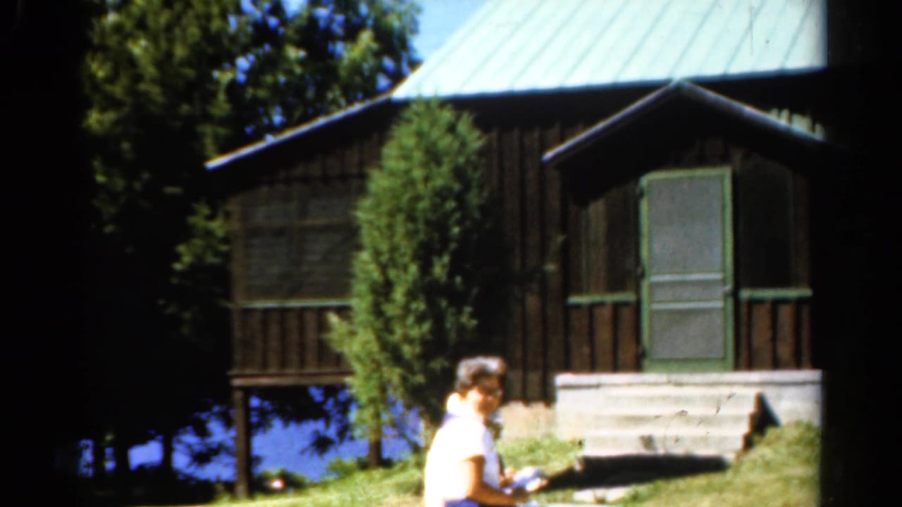 1962 Catskills frame with a woman near a rustic lakeside cabin, trees, porch steps, and blue water behind it.