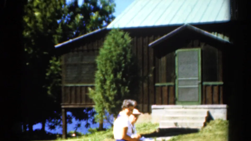 1962 Catskills home-movie frame showing two women passing in front of a wooden cabin with green trim near the lake.