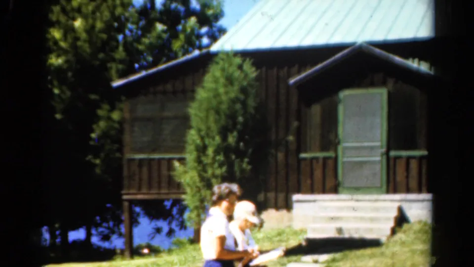 1962 Catskills frame with two women beside a lakeside cabin, one partly turned toward the boat or ground area.