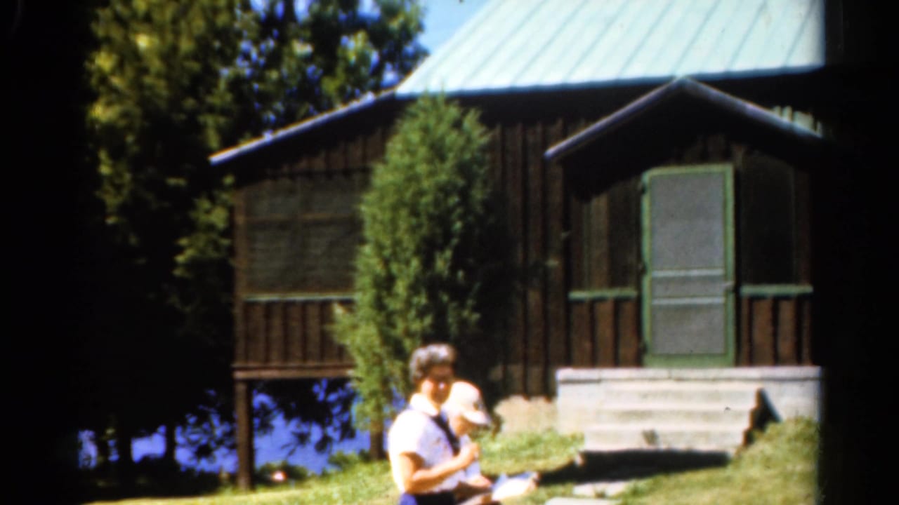 1962 Catskills home-movie frame showing adults near a lake-view cabin with dark siding, green roof, porch steps, and trees.