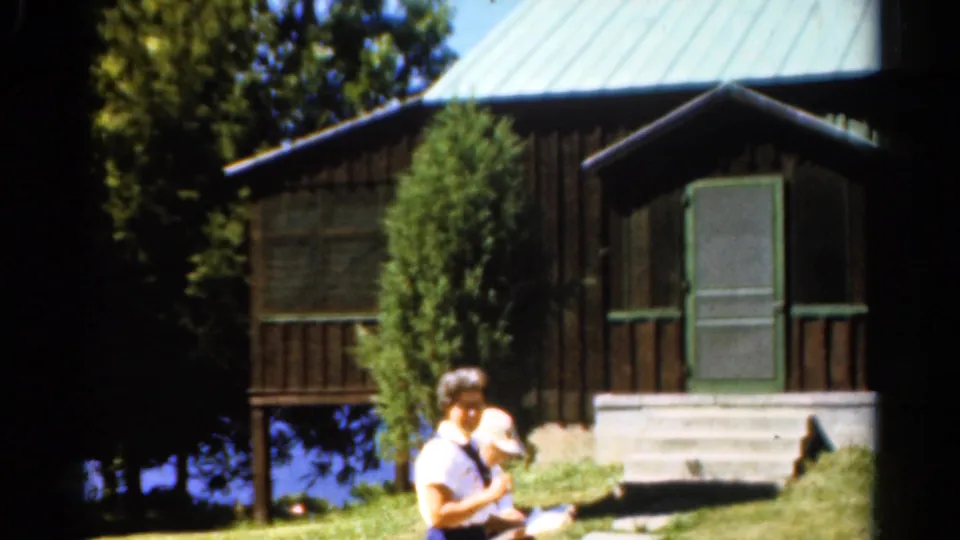 1962 Catskills home-movie frame showing adults near a lake-view cabin with dark siding, green roof, porch steps, and trees.