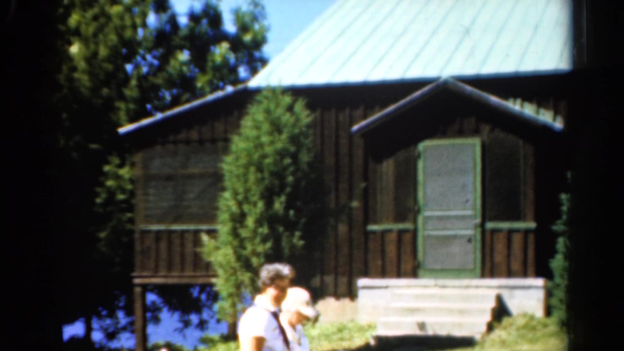 1962 Catskills frame with adults partly visible before a dark wood cabin, green roof, porch, trees, and a glimpse of lake water.