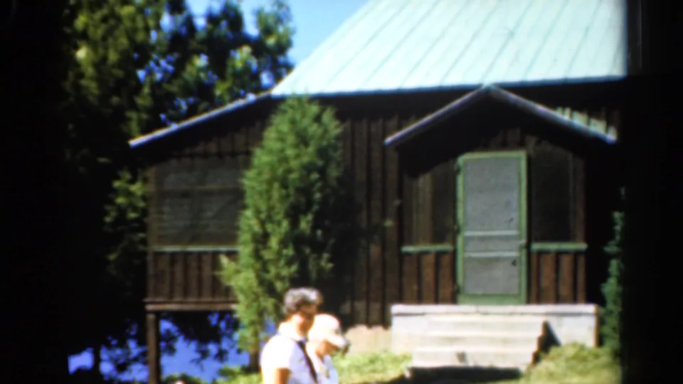 1962 Catskills frame with adults partly visible before a dark wood cabin, green roof, porch, trees, and a glimpse of lake water.