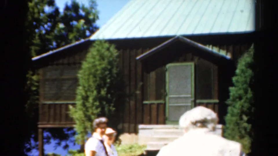 1962 Catskills home-movie frame with adults crossing the foreground near a rustic lake cabin, porch steps, trees, and water beyond.