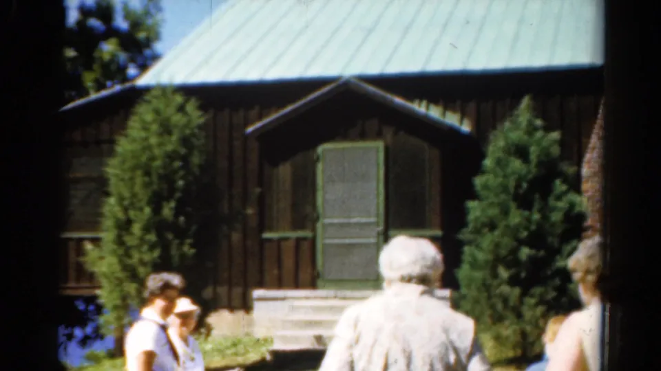 1962 Catskills frame showing adults gathered near a dark wood lake cabin with green roof, porch steps, trees, and sunlit lawn.