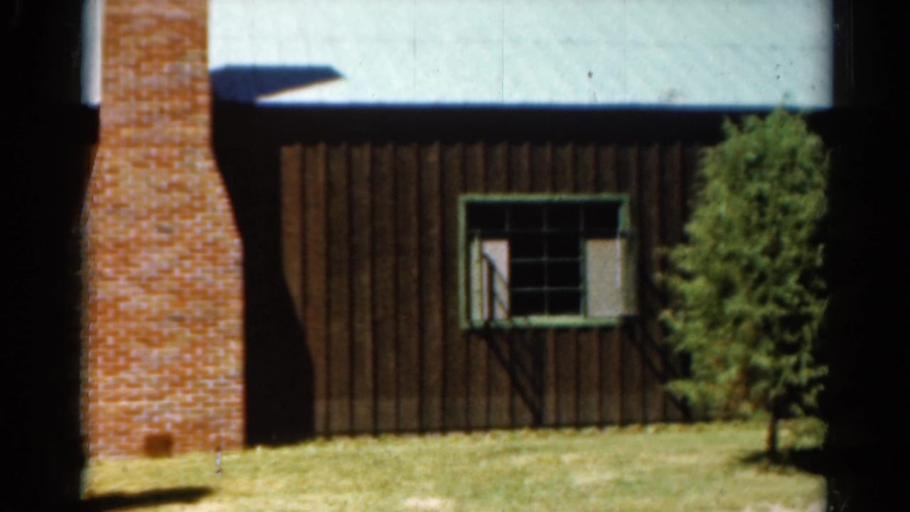 1962 Catskill-region 8mm frame showing a brown wood building, brick chimney, green-trimmed window, lawn, and small evergreen.