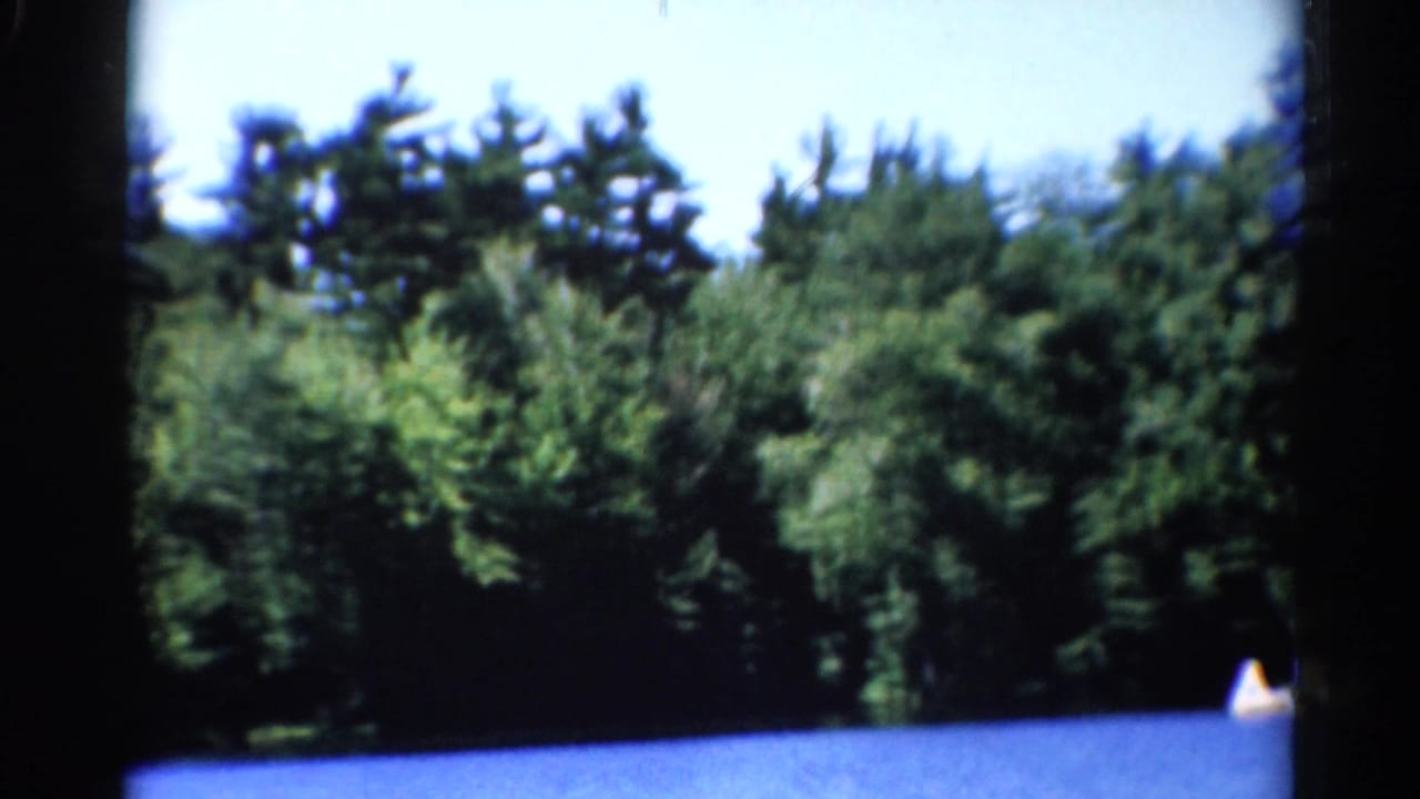 1962 Catskills home-movie frame showing a forested shoreline, blue lake water, and a small distant boat at the edge of the frame.