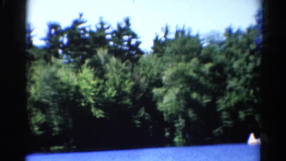 1962 Catskills home-movie frame showing a forested shoreline, blue lake water, and a small distant boat at the edge of the frame.