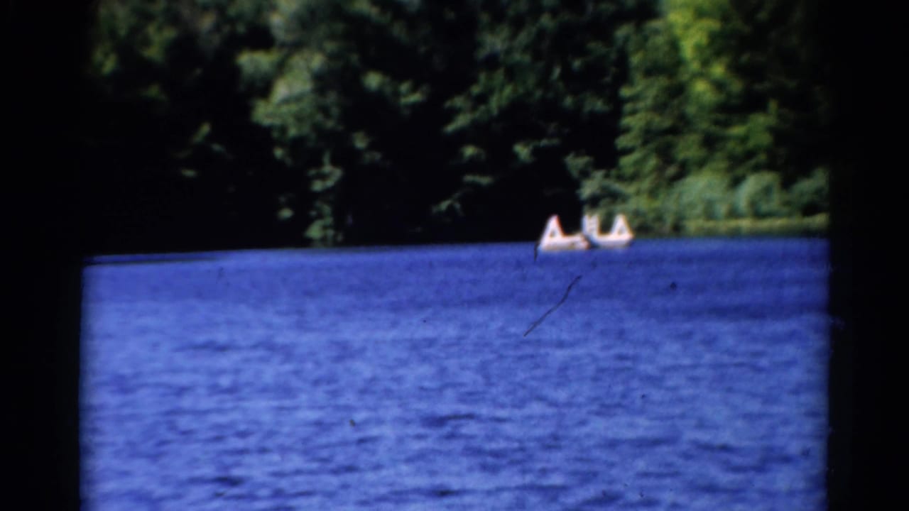 1962 Catskills lake scene with rippled blue water, dense trees, and two small boats carrying people in the distance.