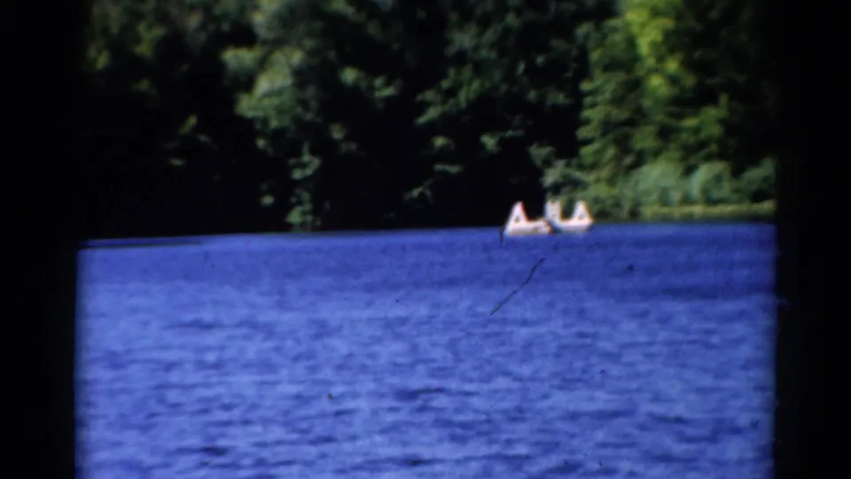 1962 Catskills lake scene with rippled blue water, dense trees, and two small boats carrying people in the distance.