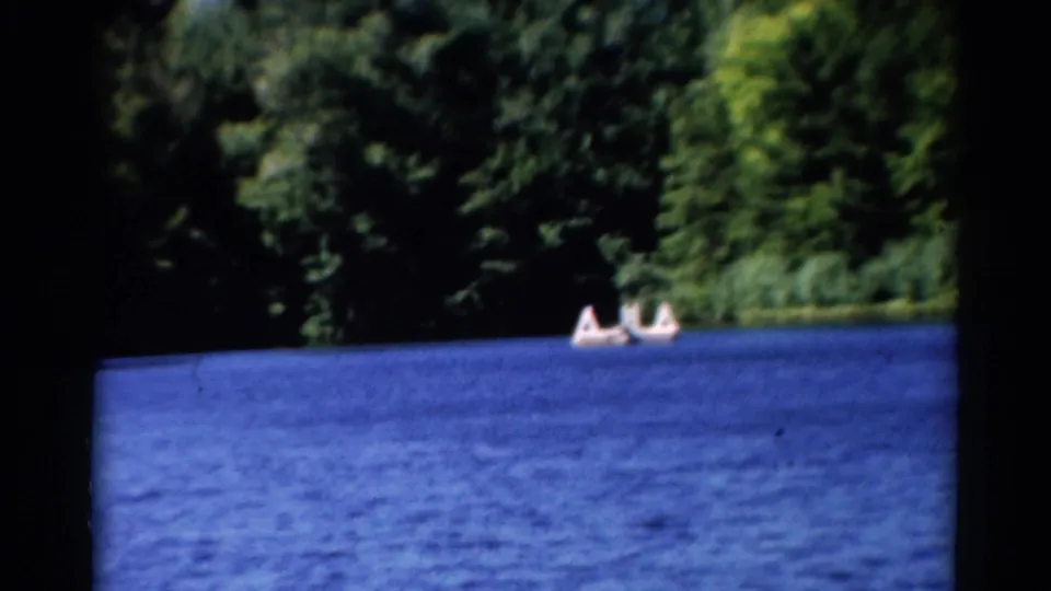 1962 Catskills home-movie frame showing people in a small boat crossing blue water near a wooded shoreline.