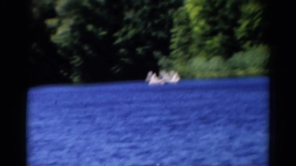 1962 Catskills frame with distant people in small boats on a blue lake, backed by a soft green wooded shoreline.
