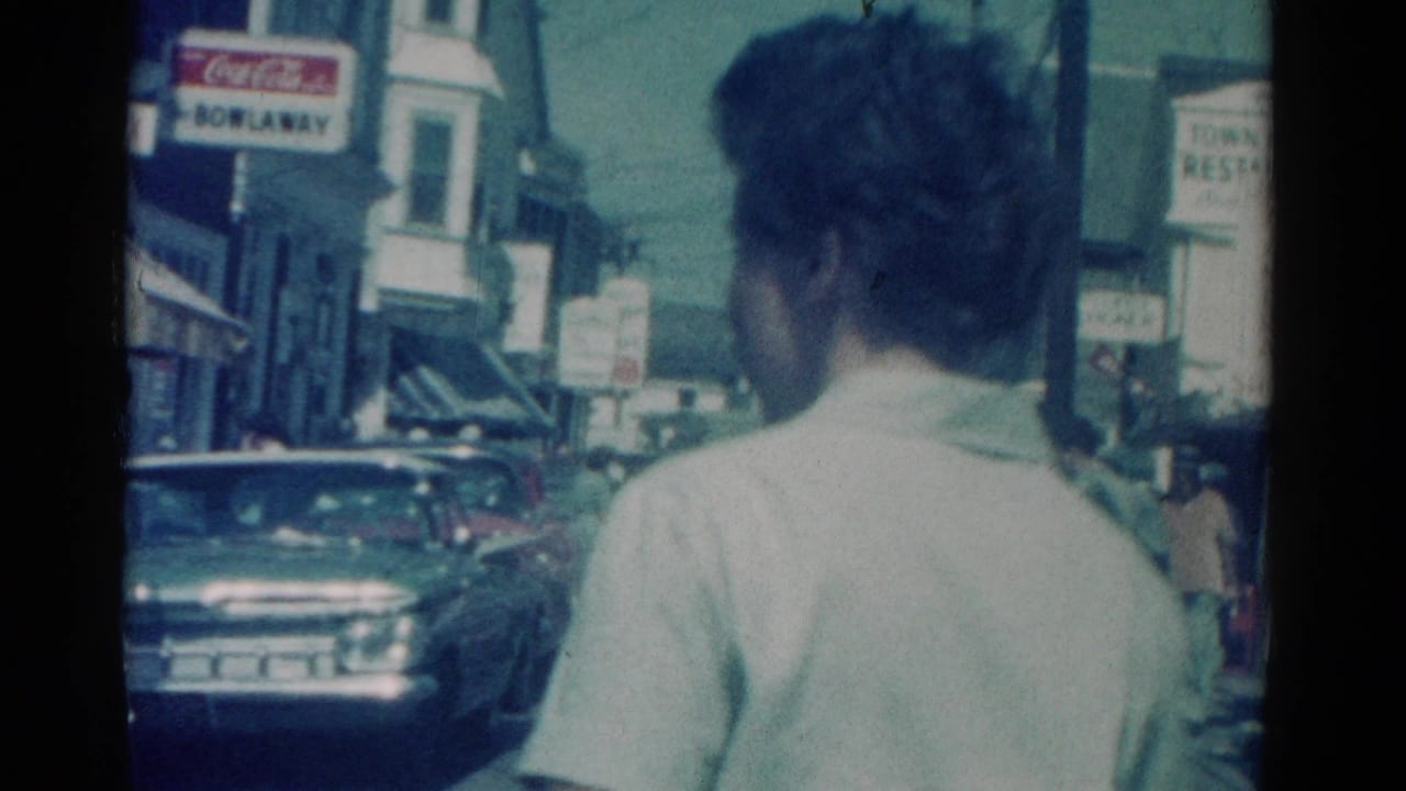 1962 Catskill, New York street view with a man seen from behind, storefront signs, parked cars, and pedestrians in the distance.