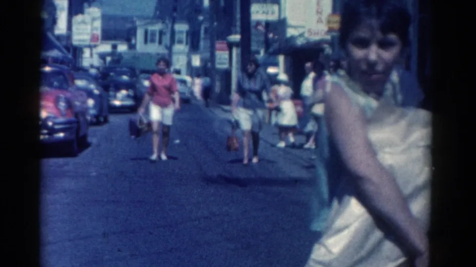 Catskill, New York, 1962 home-movie frame with a woman passing close to camera and pedestrians farther down the commercial street.