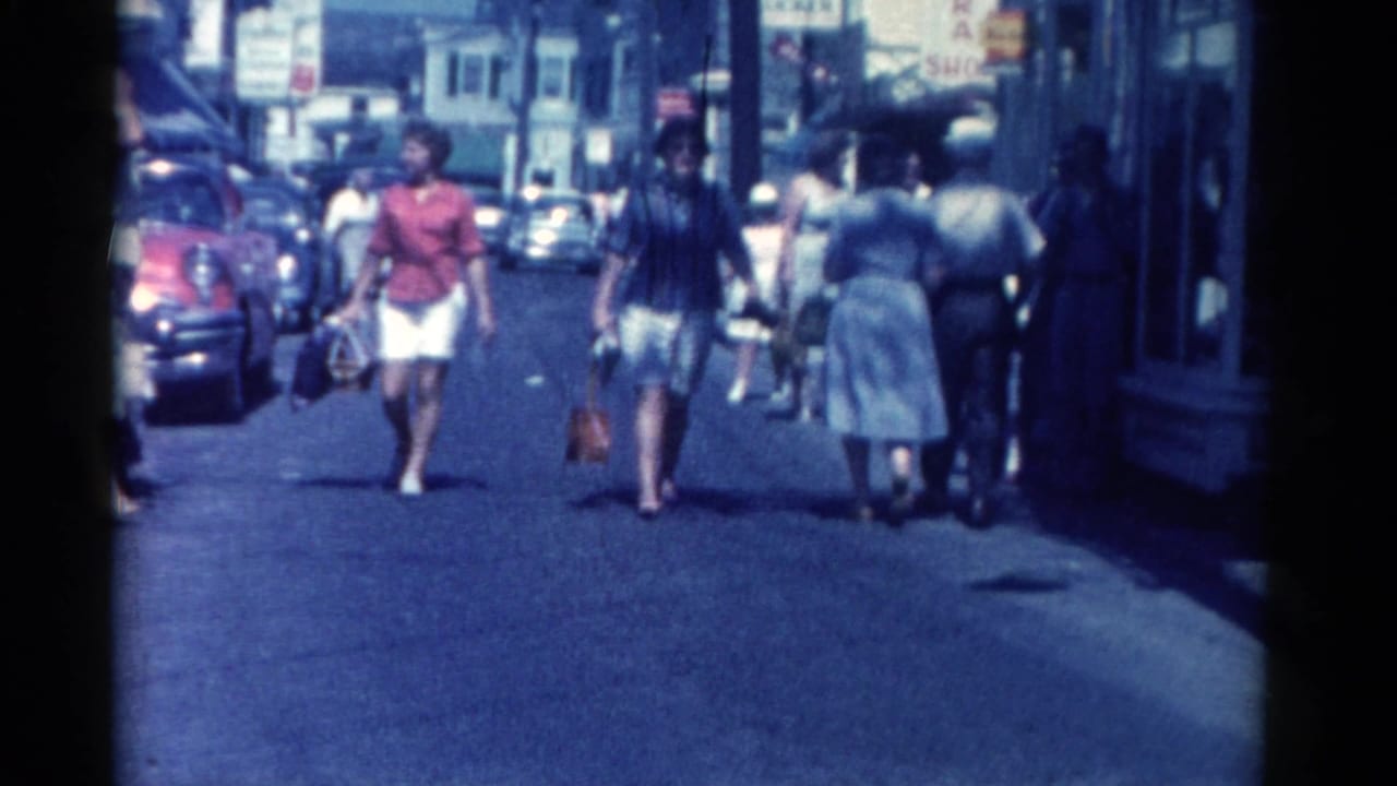 Women carrying bags walk toward the camera while other pedestrians pass storefronts.