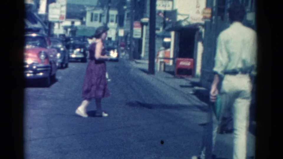 1962 Catskill downtown view with a woman crossing the street, a man near the curb, storefront signage, and parked classic cars.