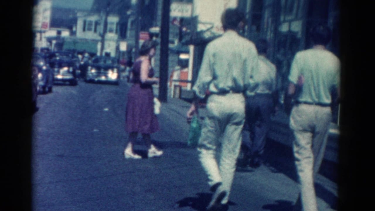 Pedestrians walk along a sunny downtown sidewalk beside storefront windows and street traffic.
