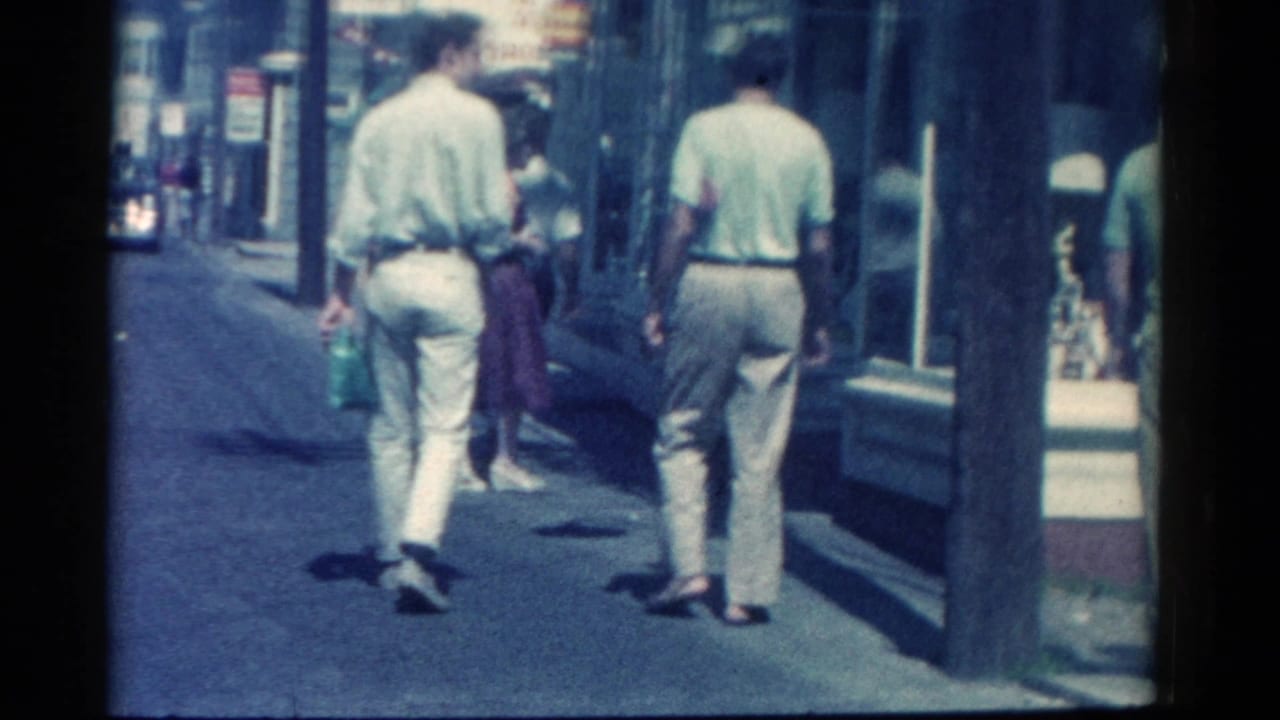 Men in light clothing walk along a Catskill retail sidewalk in 1962, with shop windows and a passing woman ahead.
