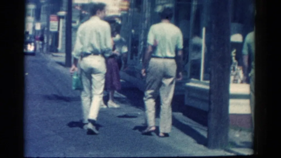 Men in light clothing walk along a Catskill retail sidewalk in 1962, with shop windows and a passing woman ahead.