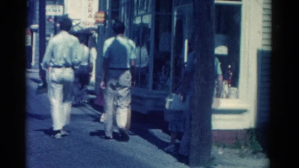 1962 Catskill main street sidewalk view with several pedestrians moving past retail display windows and street poles.