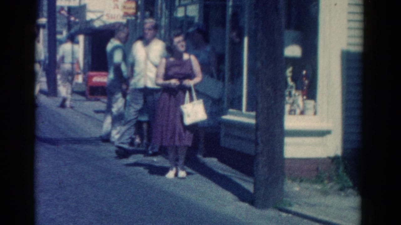 Pedestrians walk along a sunny sidewalk beside storefront windows.