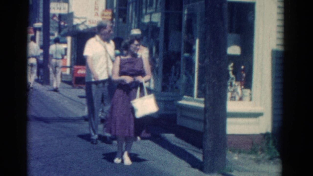 A man and woman walk together near shop windows and a utility pole.