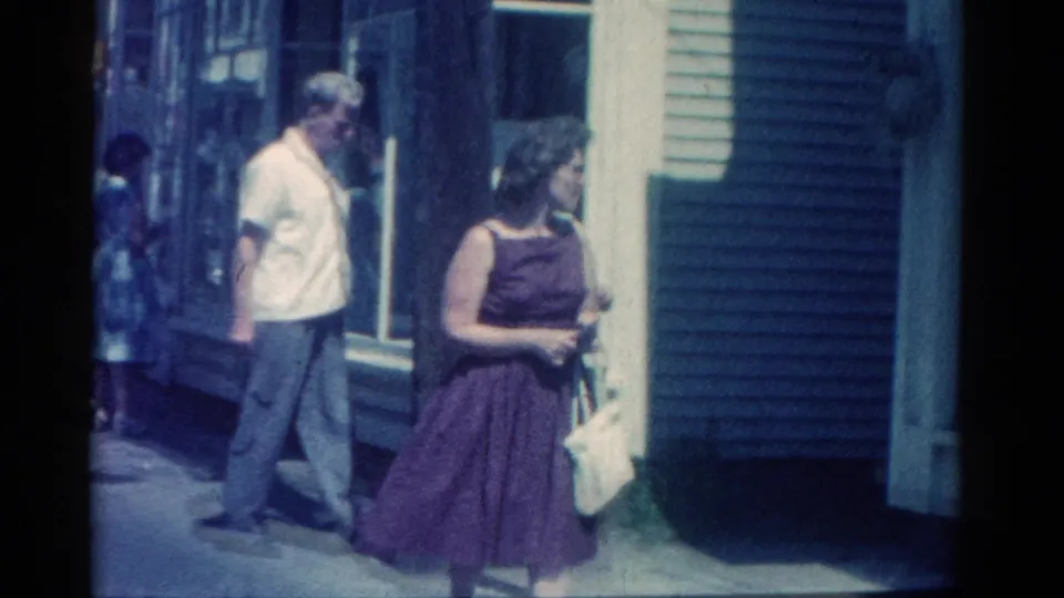 The woman in a purple dress continues along the 1962 Catskill sidewalk, with storefront siding and a man behind her.